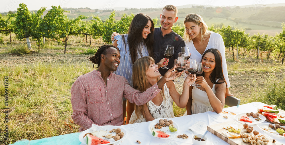 Naklejka premium Happy friends having fun drinking red wine and eating together - Multiracial people doing party at summer time in countryside with vineyard in background - Main focus on people faces