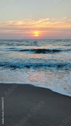Vertical slow motion sunrise scenery with sun glowing through clouds above sea waves and sandy shoreline