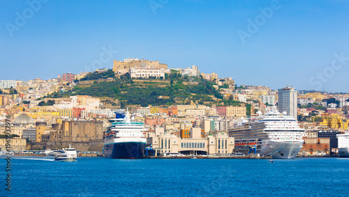 Panoramic view of Naples Italy coastline with harbor, cruise ships and historic buildings. Naples features Castel Sant Elmo on hill above urban architecture. Clear weather, calm sea, bright daylight 