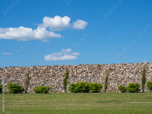 Minimalist landscape with a stone embankment wall, green grass, and small shrubs under a bright blue sky with scattered white clouds. Clean, natural outdoor scene with copy space, symbolizing calm
