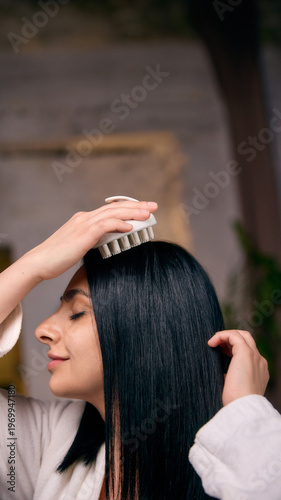Woman massaging scalp with brush during hair care routine. Concept of hair treatment process, relaxation ritual, scalp stimulation, healthy hair growth, self care beauty routine.