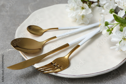 Beautiful table setting with golden cutlery and blooming branch on grey background, closeup
