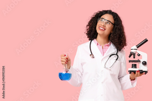 Female African-American doctor with microscope and flask on pink background