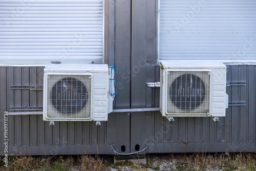 Two outdoor air conditioning units fixed on a grey modular building wall, beneath closed white roller shutters, illustrating essential climate control technology