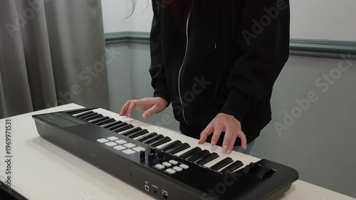White woman in hoodie practices precise finger technique on electronic keyboard closeup, highlighting scales, arpeggio patterns and hand posture against neutral backdrop