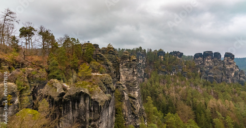 Large panorama of the jagged Bastei rock formations, including the Wehltürme and the Ferdinandstein lookout, where visitors admire the Saxon Switzerland landscape on a cloudy winter day.