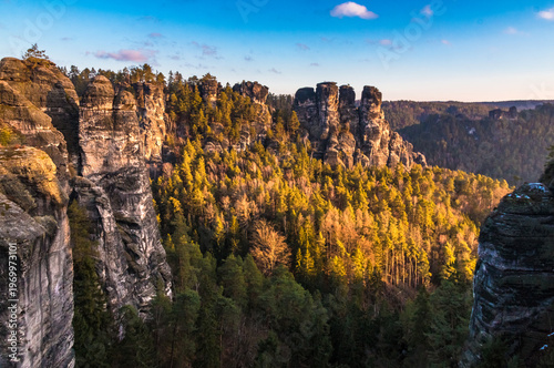 Picturesque panoramic view of the jagged Bastei rock formation in Germany’s Elbe Sandstone Mountains, a landmark of Saxon Switzerland, glowing in golden winter sunrise light under a blue sky.