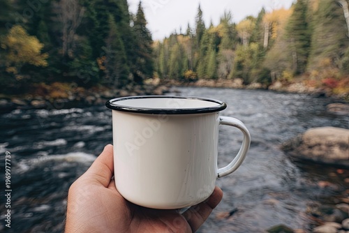 Close-up of a hand holding a white metal mug against a scenic river and forest