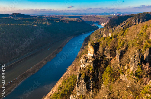 Downstream view along the Elbe River toward the Wartturm, the collapsed sandstone tower, in a picturesque panoramic winter scene with blue sky and golden sunrise light above the Bastei cliffs.