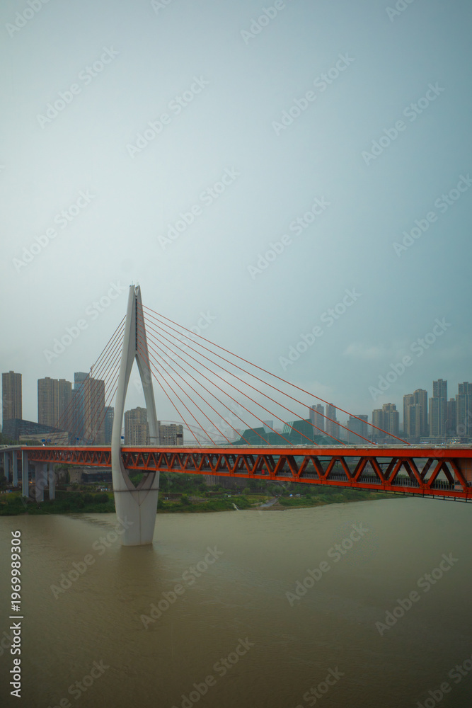 Obraz premium Red bridge spans wide river under cloudy sky. Modern structure rises from calm, muddy waters. City skyline fades softly in hazy background. Steel cables stretch from tall central pylon