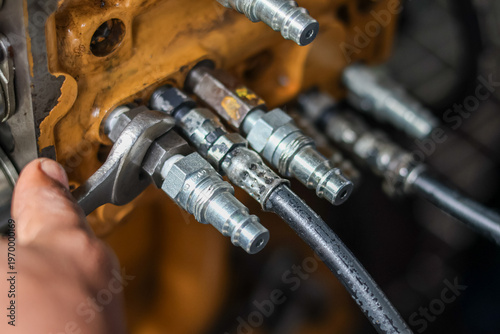 Close-up of a mechanic hand using a wrench to tighten hydraulic hose fittings on a yellow industrial engine block.
