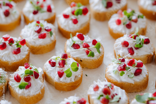 A close-up of appetizers featuring toasted bread slices topped with cream cheese, pomegranate seeds, and microgreens, arranged on a neutral surface.