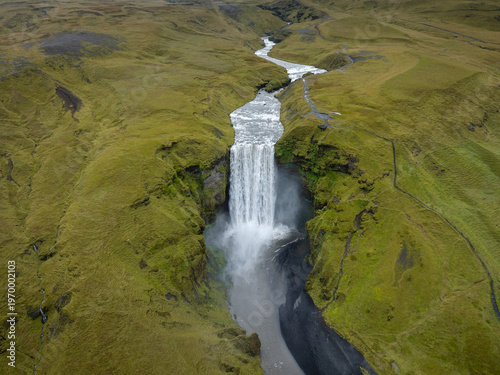 Aerial view of Skogafoss waterfall cascading powerfully through the verdant Icelandic landscape, contrasting with the dark, rocky terrain below, Skogafoss, Rangarthing eystra, Iceland.
