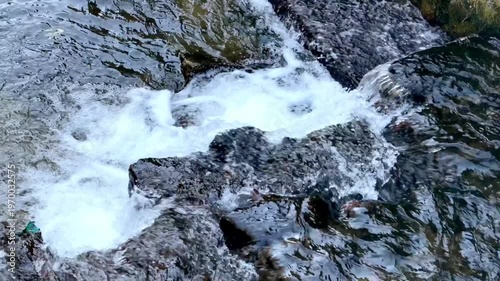 Vibrant natural stream water vigorously cascades over dark rugged rocks, producing brilliant white foamy rapids and clear ripples, conveying nature's dynamic power and serene environmental essence