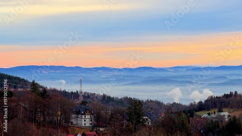 Misty mountain vista shows blue hills, village homes, communication tower, and smoke at dawn, illustrating nature and human impact