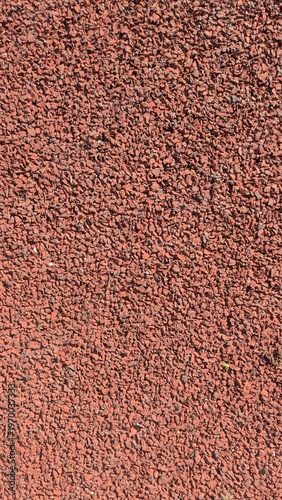 Close-up of a textured red rubber surface, commonly used in athletic tracks and playgrounds, showcasing its granular composition under natural light.