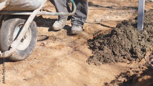 Laborer Transporting Cement Mixture Using Wheelbarrow During Construction. Worker Navigating Construction Area With Wheelbarrow Filled With Mortar Surrounded By Dirt And Supplies