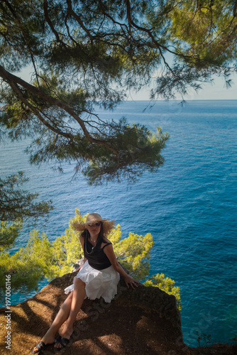 Girl in summer skirt and straw hat dancing on cliff edge with skirt flowing over boulders waves of Adriatic Sea and green Montenegro hills in distance