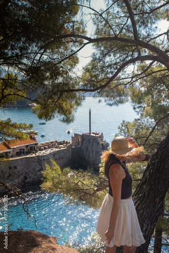 Woman in summer skirt and straw hat spins with open arms on cliff above rocks Adriatic sea and green hills Montenegro travel inspiration scene