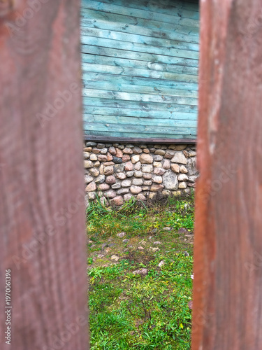 Creative narrow perspective through wooden fence slats reveals rustic turquoise timber wall above stone foundation and green grass within rural backyard.