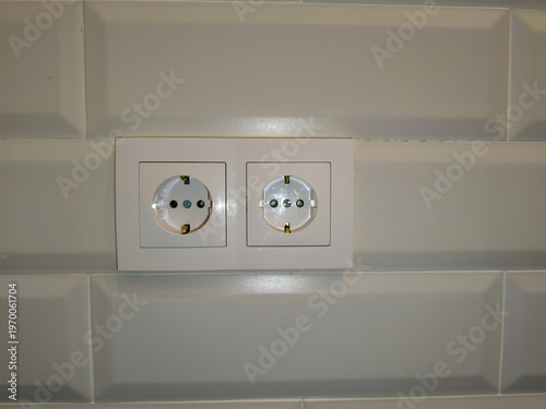 White dual electrical outlet mounted horizontally on beveled subway tile kitchen backsplash background providing domestic energy supply connection points for household appliances