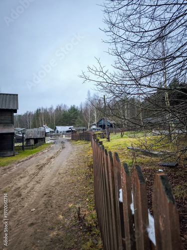 Rural dirt path winds through traditional village featuring dark weathered log buildings and long wooden picket fence under gloomy overcast sky near forest edge.