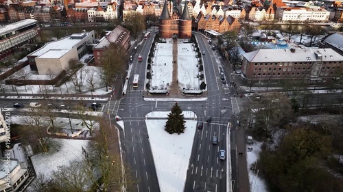 Drone pull-out footage from decorated Christmas tree placed on a snow-covered traffic island reveals the Holstentor, snowy streets and historic skyline of Lübeck.