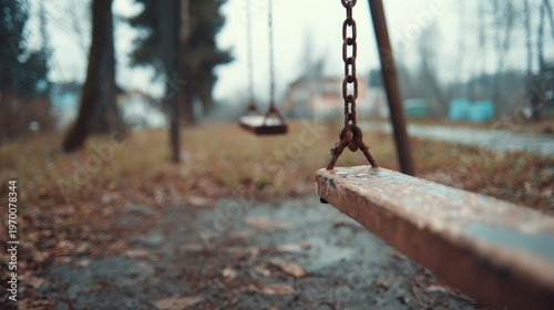 Empty wet wooden swing set with rusty metal chains in an outdoor park on a gloomy autumn day surrounded by fallen leaves and blurred trees