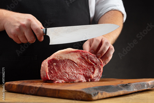 Male hands cutting raw beef meat. Male hands of Professional butcher, chef holding a large knife and cutting a piece of raw meat on a cutting wooden board. 