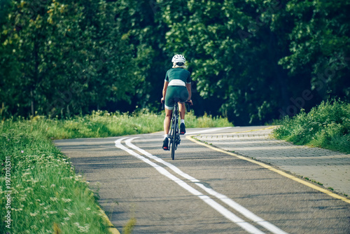 A woman cyclist in green gear rides away along a path surrounded by flowering trees, captured from behind at a wider distance. The image conveys freedom, exploration, and the beauty of summer.