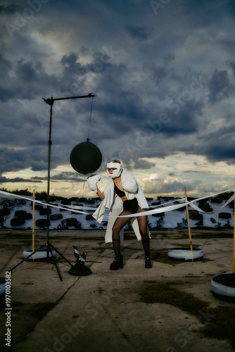 Young athletic woman wearing white boxing gloves and protective helmet strikes a punching bag inside a conceptual outdoor ring.