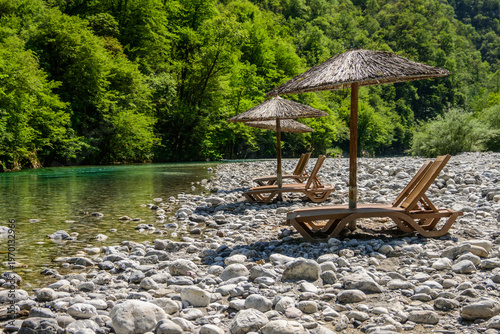 Rustic straw umbrellas and wooden loungers are set on a rocky riverbank beside the transparent turquoise Shala River, with dense green mountains as a backdrop in Albania