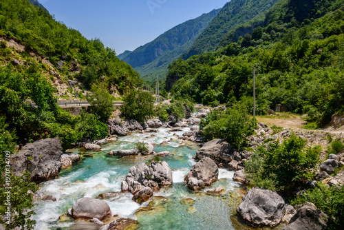 A vibrant Valbona Valley river with stunning turquoise water cascades over rocks, surrounded by densely forested mountains under a clear blue sky, evoking wilderness in Albania.