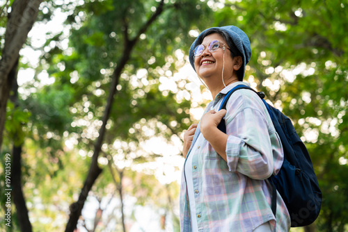 Portrait of Asian mature woman in a hat with backpack behind her back, an Asia active senior woman enjoying nature in autumn park. Standing on a trail in a forest outdoors. Enjoying active travel trip