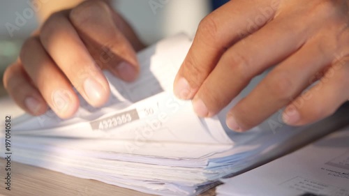 Hands gently flip through a stack of papers. Natural light illuminates the workspace. Fingers show care as documents are handled. Documents rest on a wooden desk surface. Hands move with precision