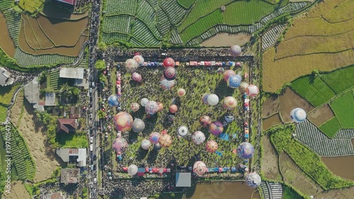 Aerial top-down view of colorful hot air balloons being prepared for launch in a crowded festival field. Surrounded by rice fields and rural landscapes, the scene captures vibrant patterns.