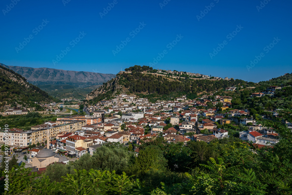 Fototapeta premium A broad, vibrant panorama of Berat, Albania, captured on a sunny day, featuring the city, the Osum River, and the forested castle hill against distant mountains.