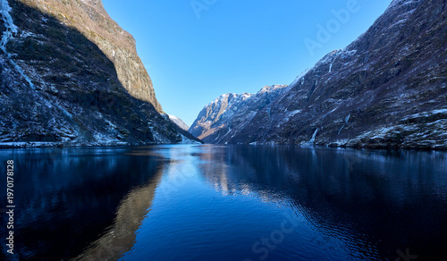 Majestic Winter Fjord Landscape: Steep Snow-Capped Mountains and Serene Water Reflection under Clear Blue Sky in Norway