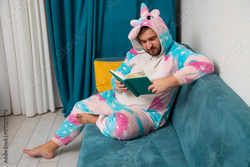 Horizontal photo. Young adult white man with beard wearing pink unicorn costume sits on sofa in living room and reads blue book. Concept of learn, rent apartment, leisure time at home, relax, rest