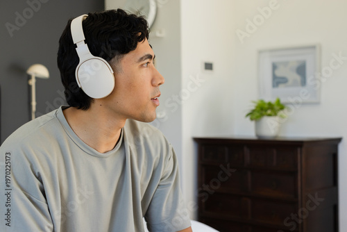 Asian man sitting on left wearing white headphones and grey T-shirt, speaking near lamp and clock