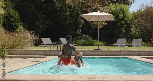 Father lifting son on shoulders in backyard pool, pushing into splash while playing near beach ball