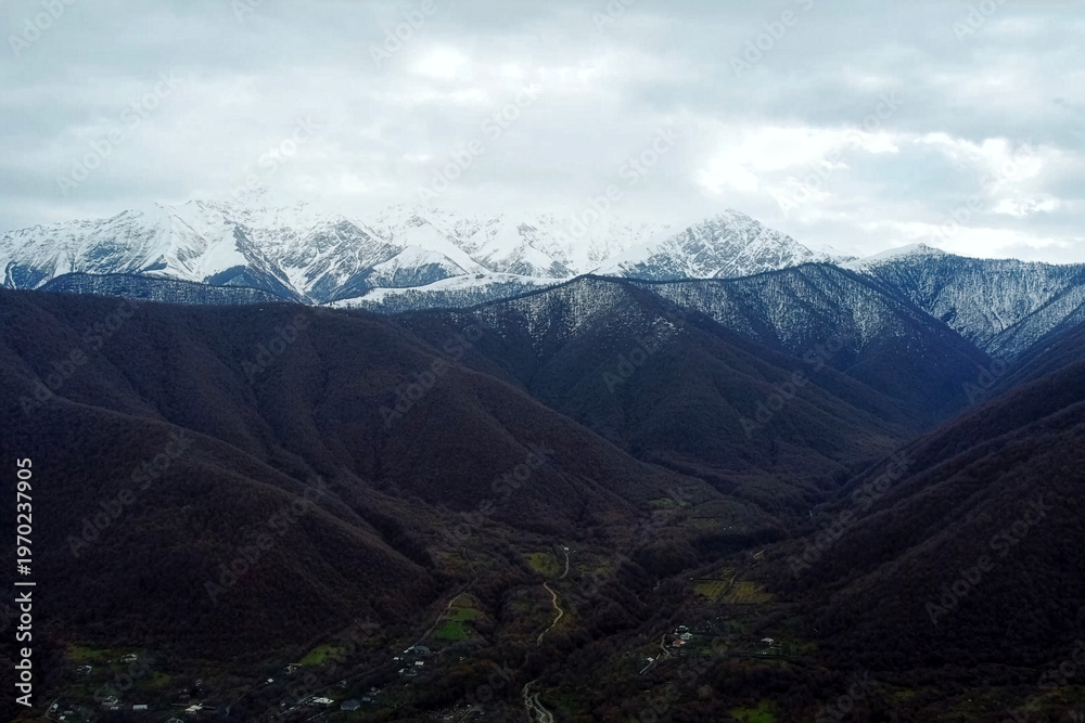 Fototapeta premium Aerial View of Snow-Capped Mountain Range Over Small Rural Village. A scenic landscape showing towering, snow-covered peaks overlooking a valley with a small town and a dry riverbed