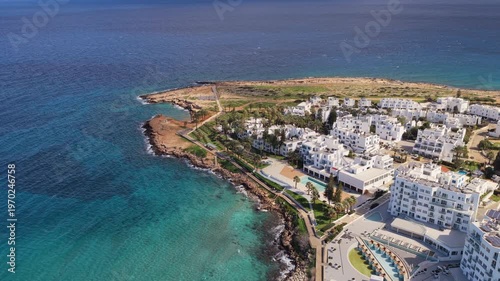 Aerial drone view of a coastal resort area with white buildings, palm trees, and clear blue sea along a rocky shoreline in Cyprus
