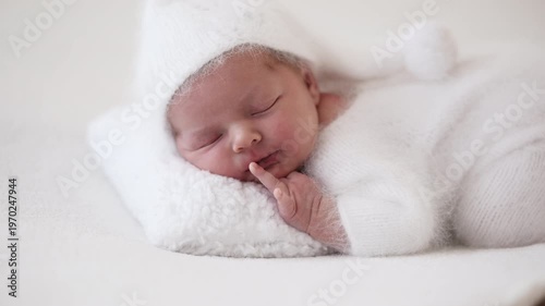 Newborn Sleeps Quietly On Fluffy White Pillow Wearing Cozy White Outfit And Hat With Hands Tucked Under Chin. Newborn Photo Shoot.