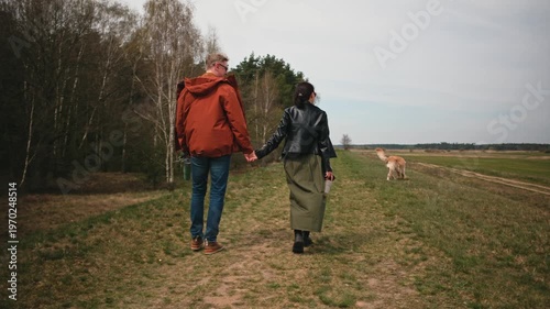 Couple walk hand in hand on a grassy trail with a golden retriever dog.