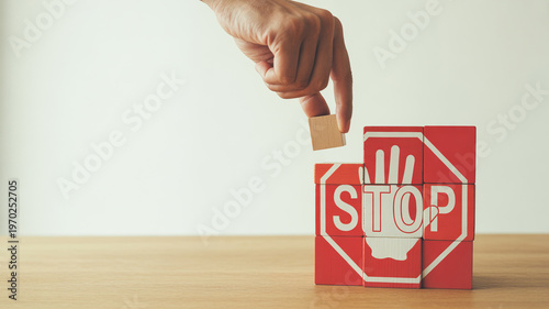 A human hand carefully places a small wooden block atop red cubes spelling the word STOP with a stop sign symbol on a clear light background creating 