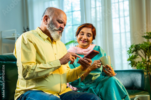 Photography Retired Asian couple sitting on a sofa while sharing a smartphone for digital tasks at home