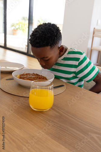 African American school-age boy leaning over white bowl at home dining table, orange juice glass
