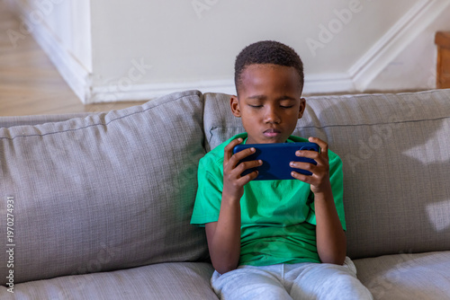 African American boy child sitting on light-gray sofa at home holding blue smartphone