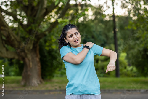 Young woman feeling severe shoulder pain and discomfort during an outdoor workout or running session, holding her painful shoulder with a suffering expression on her face